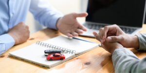 An insurance agent is discussing auto insurance policy options with a client. They are sitting at a desk and there is a computer, notebook, car keys and red toy car on the desk.