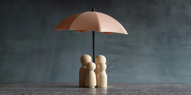 Four small wood figures shaped like people are standing under an umbrella.