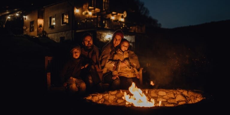 A family of four is sitting near a fire pit on a fall evening.