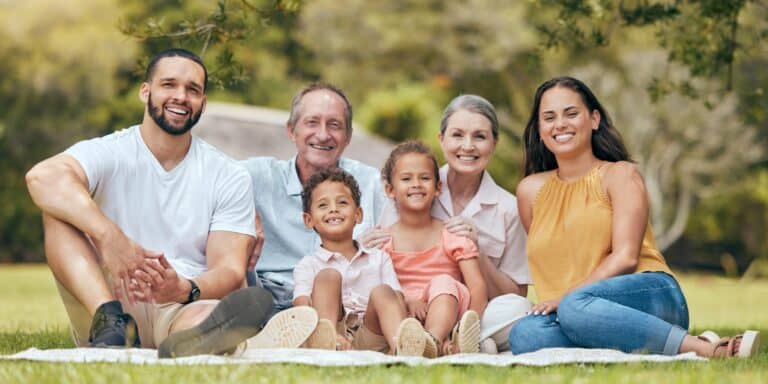 A family sitting on a blanket at a park including grandparents, parents, and two children.
