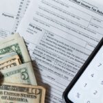 Image of a 1040 tax form, dollars, coins, and a phone calculator on a desk.
