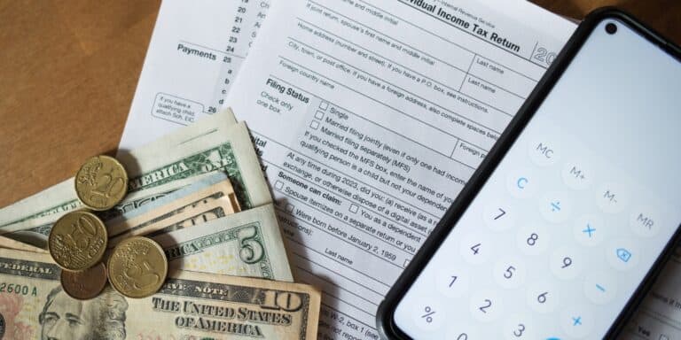 Image of a 1040 tax form, dollars, coins, and a phone calculator on a desk.