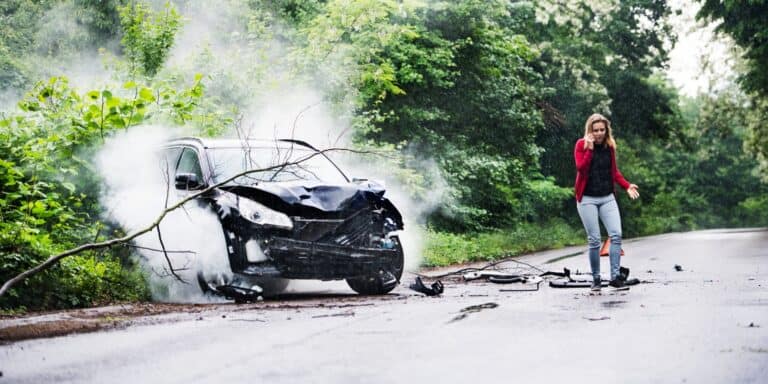 A woman is making a phone call as she walks away from her damaged vehicle after a car accident.